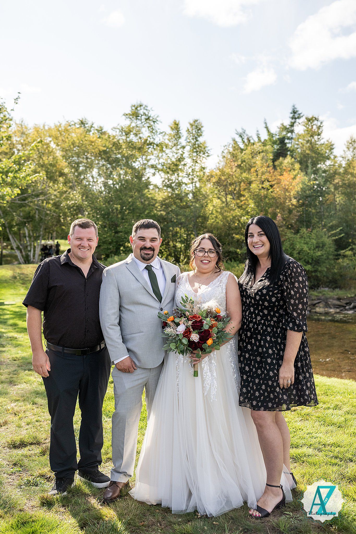 Outdoor wedding group photo with people in formal attire standing together against green scenery.