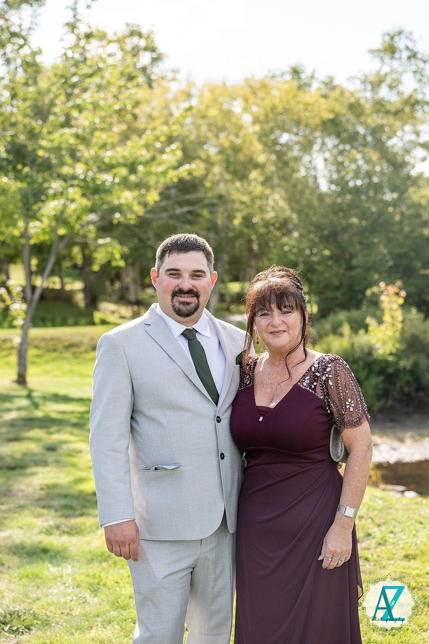 Wedding couple posing in formal attire with grey suit and burgundy dress against natural outdoor background.