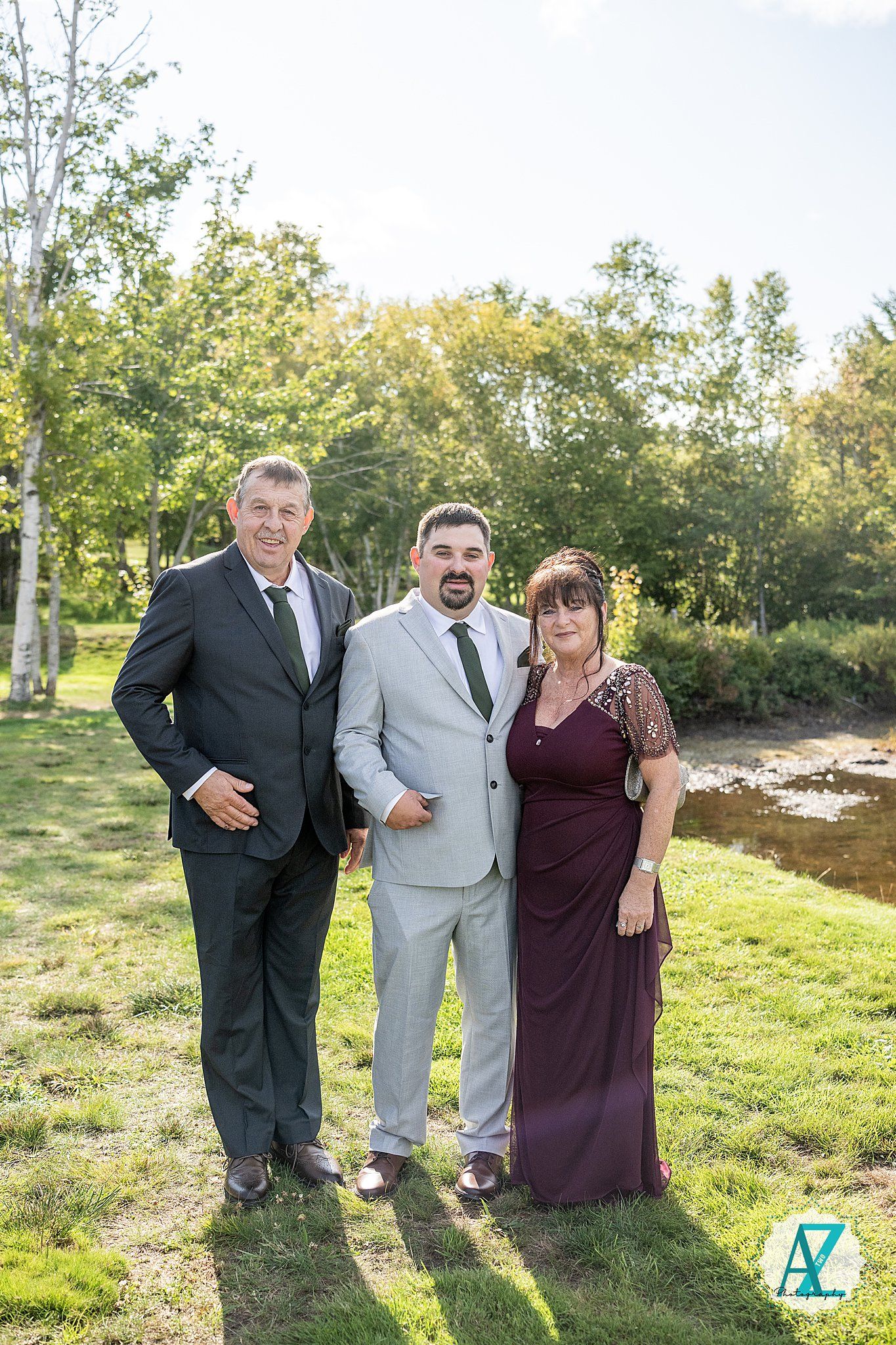 Three people in formal attire pose together on a sunny lawn with trees and a stream in the background.