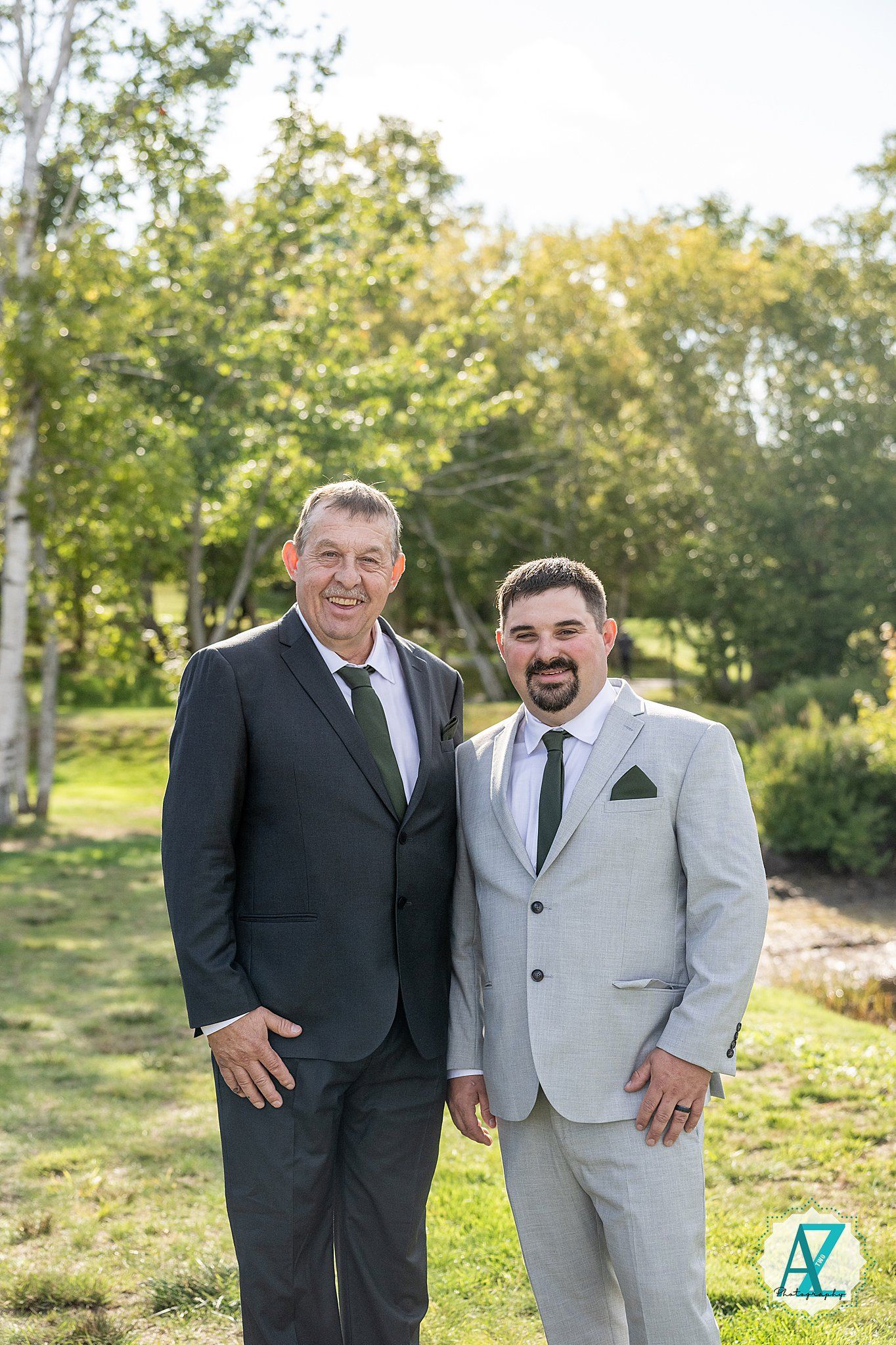Two men in formal attire stand together outdoors in a grassy area on a sunny day.