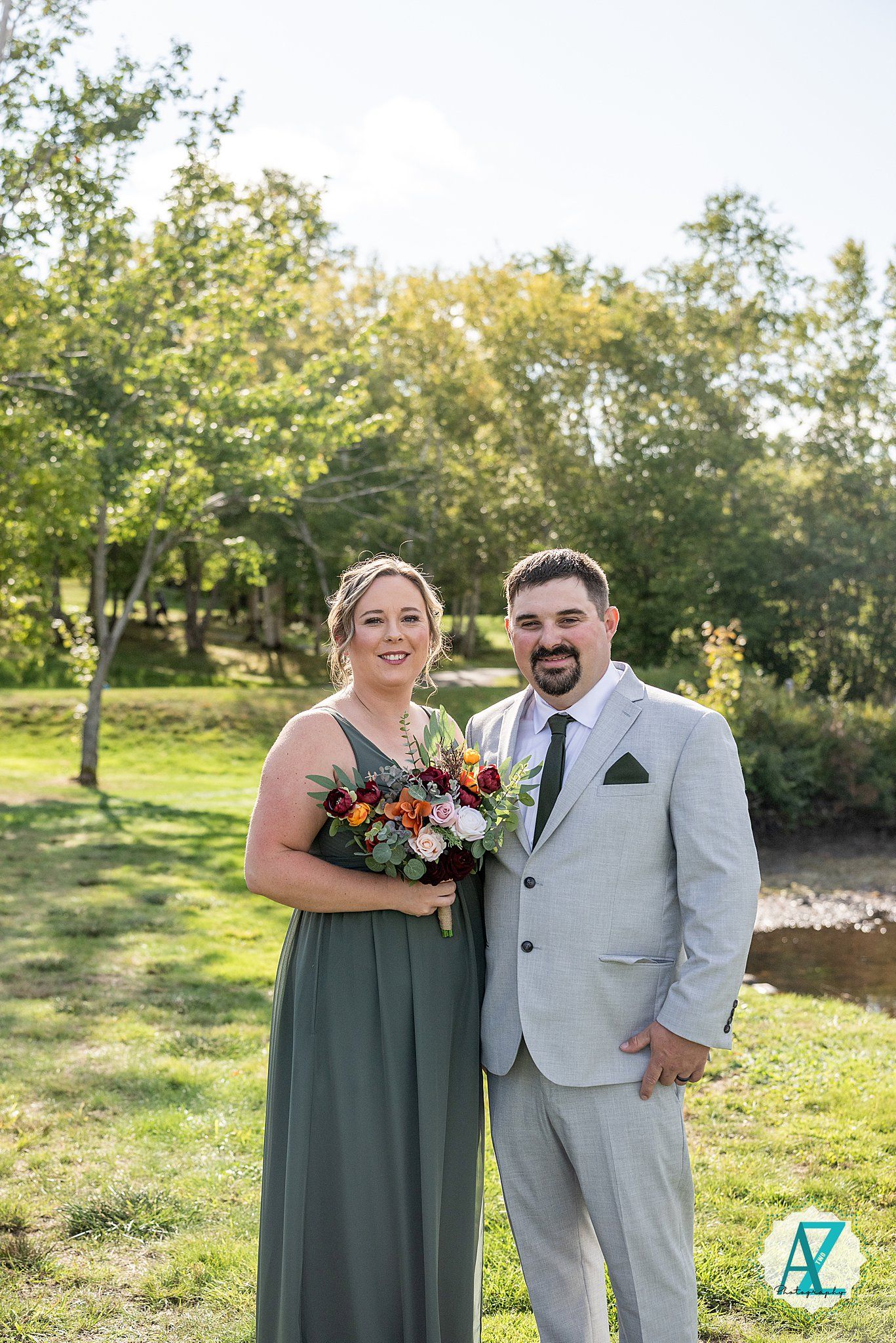 Wedding couple poses on grassy lawn wearing sage green dress and light gray suit with autumn-themed bouquet.