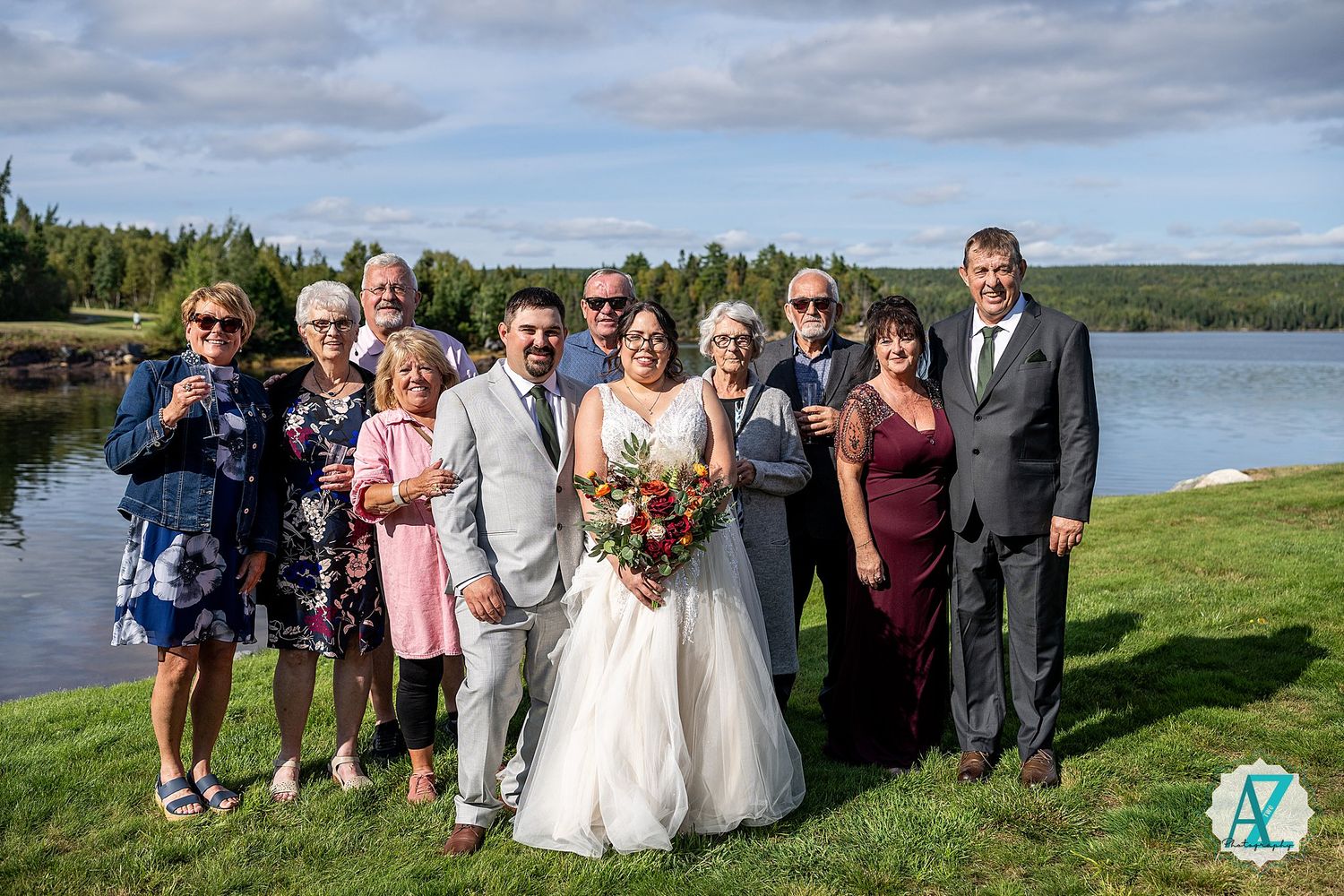Large wedding party gathers for outdoor group photo on grassy hill overlooking lake.
