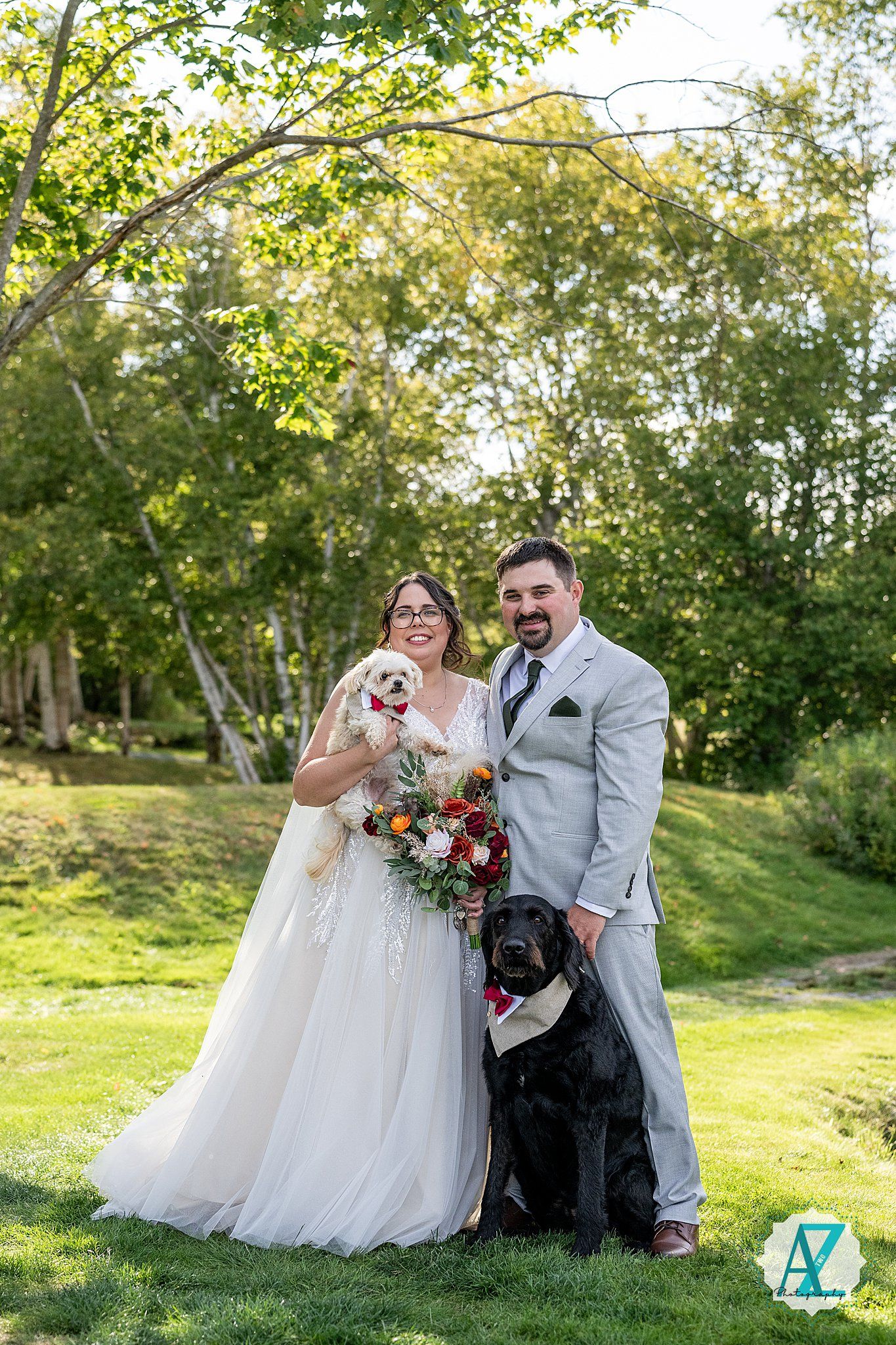 Newlyweds pose outdoors in a sun-dappled garden setting with their black Labrador dog on their wedding day.