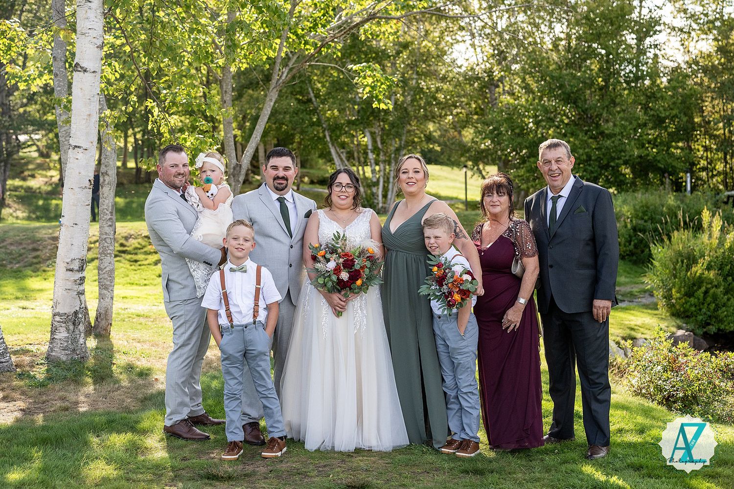 Wedding party gathers for outdoor photos in a sunlit garden setting with aspen trees and greenery in the background.