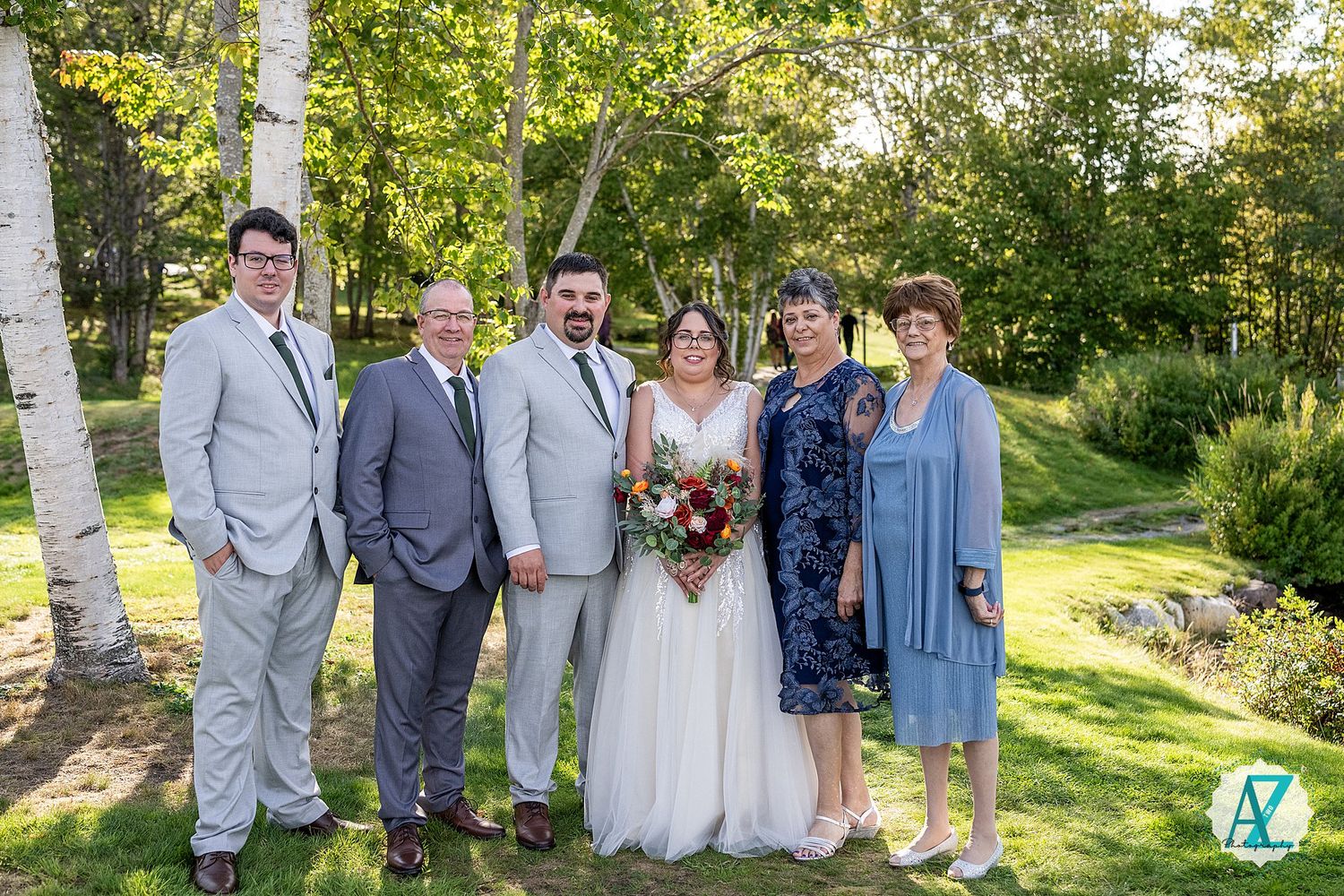 Wedding group poses in front of white birch trees and lush landscaping on a bright sunny day.