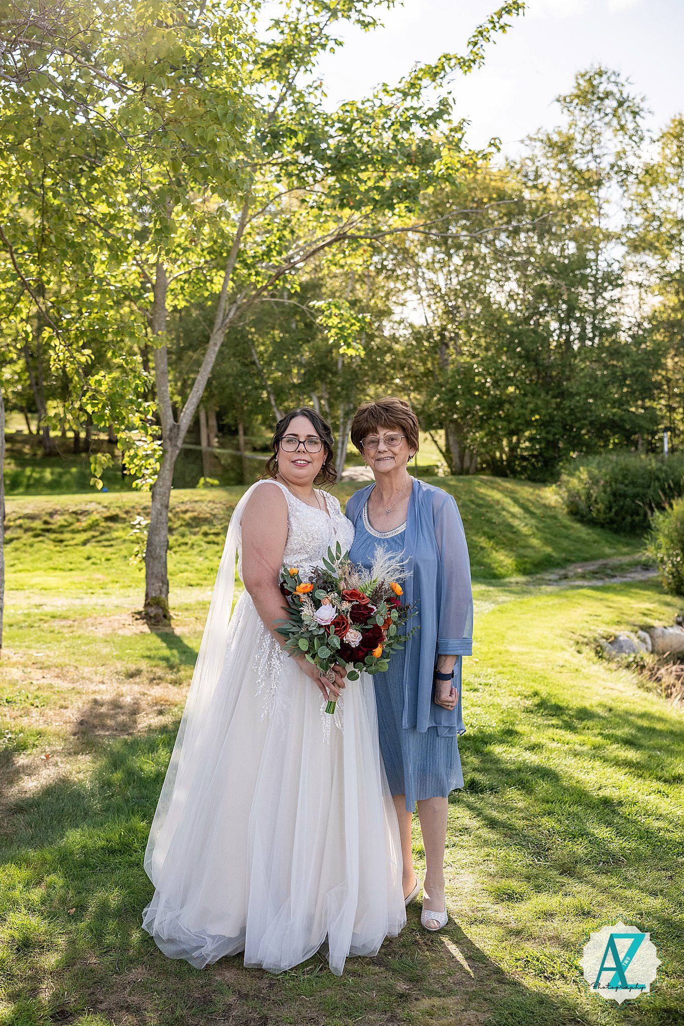 Two people pose in a sunny garden setting, one wearing a white wedding dress holding a colorful floral bouquet.