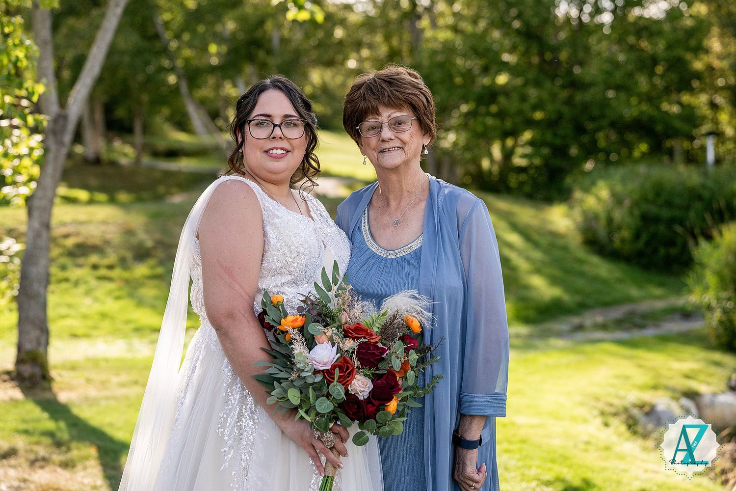 A pair stand together outdoors on a sunny day, with one wearing a bridal gown and holding vibrant flowers.