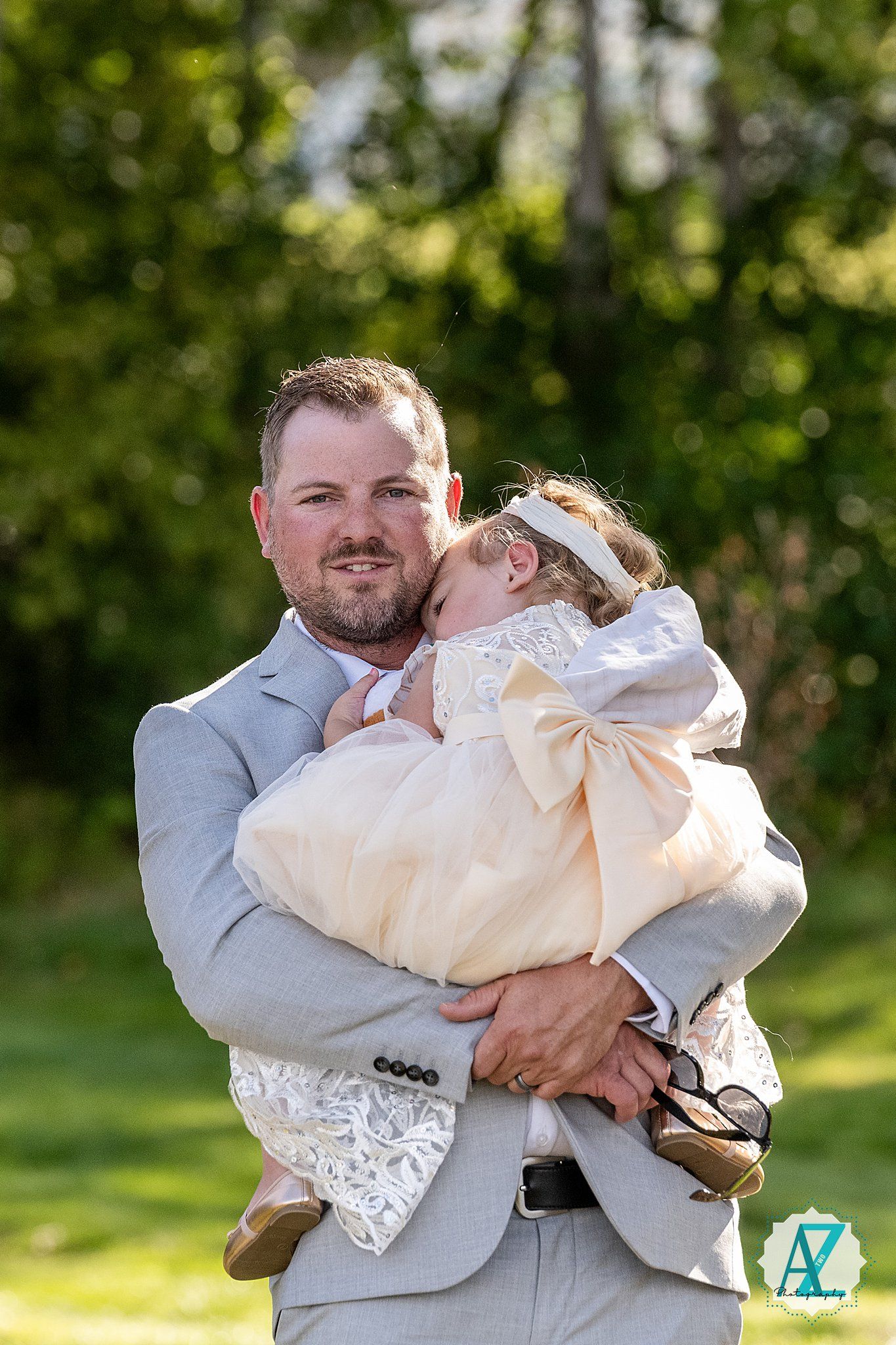 A heartwarming embrace between a man in a gray suit and a small child in white unfolds in a sun-dappled garden setting.