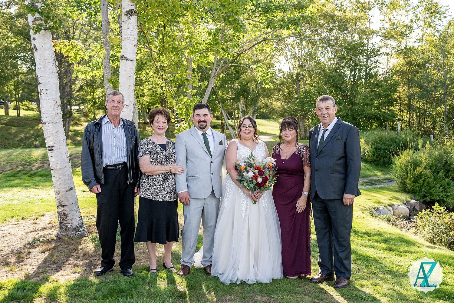 A wedding party poses outdoors between white birch trees on a sunny summer day in a garden setting.