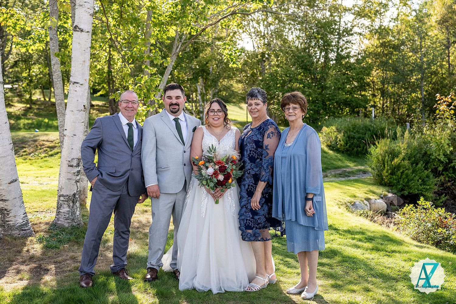 A wedding party poses together outdoors on a sunny day with birch trees and greenery in the background.