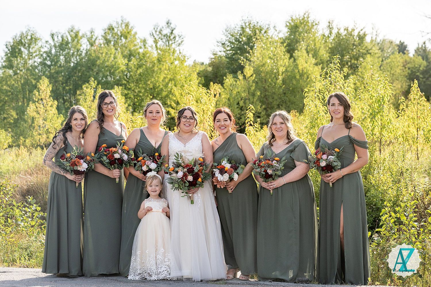 Wedding party stands together in sage green dresses and white gowns against a golden sunset background.
