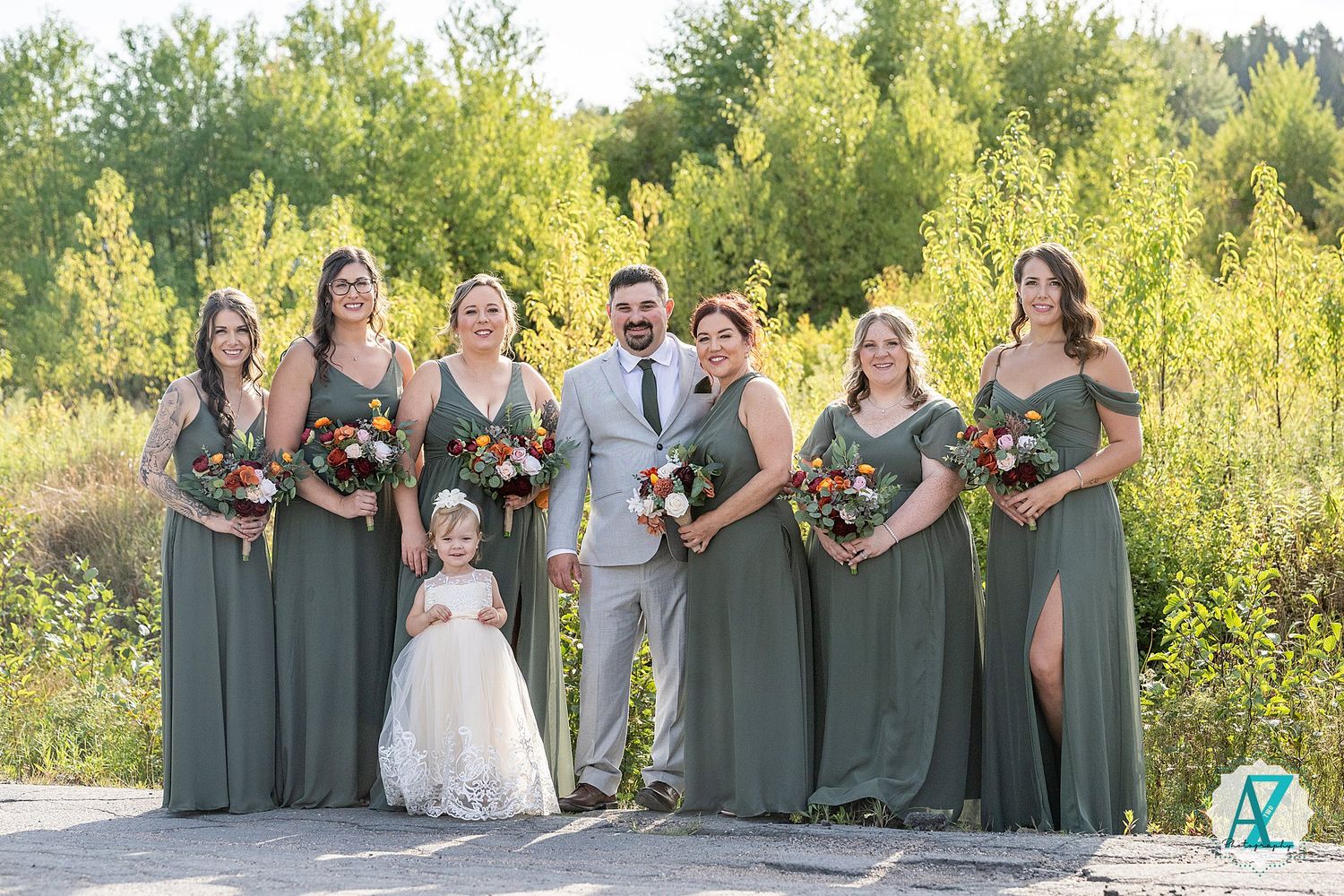 A wedding party wearing sage green dresses and gray suits poses together in a sunny outdoor field setting.