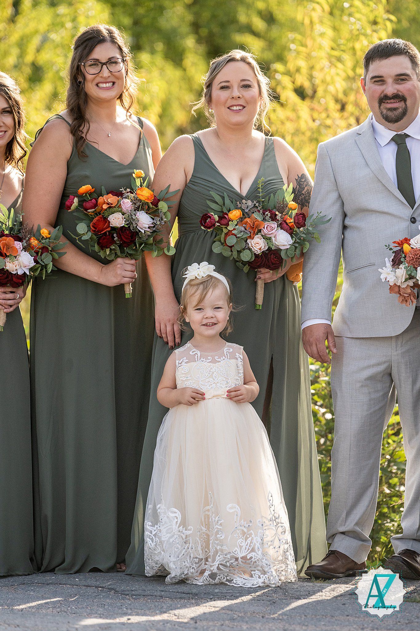 Wedding party members in sage green dresses and grey suit pose outdoors with a flower girl in white lace dress.