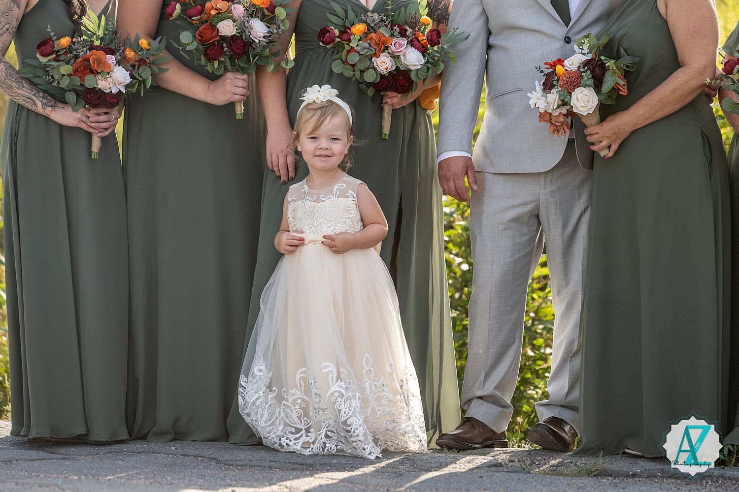 Wedding party members in sage green dresses hold vibrant orange and white floral bouquets during an outdoor ceremony.