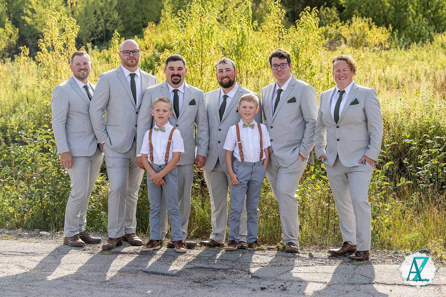 Wedding party in light gray suits and suspenders poses outdoors against a natural backdrop of yellow wildflowers.