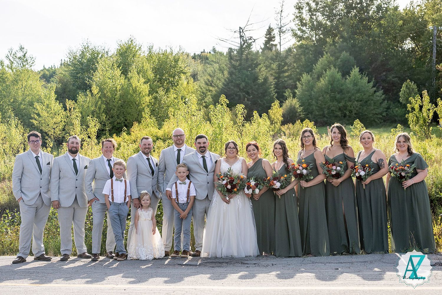 A wedding party poses in green and grey attire for a group photo outdoors on a sunny summer day.
