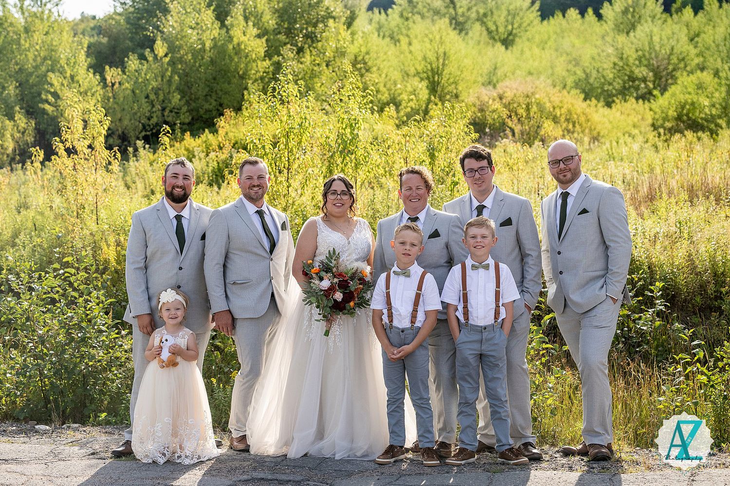 Bridal party in gray attire stands together in front of lush yellow summer foliage.