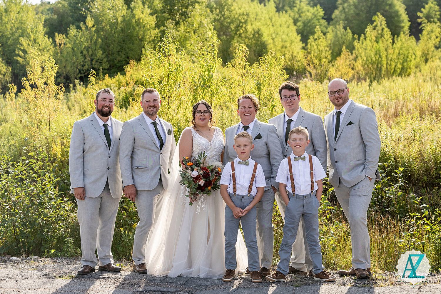 Wedding party in light gray suits and white dress pose together on a path with golden summer field in background.