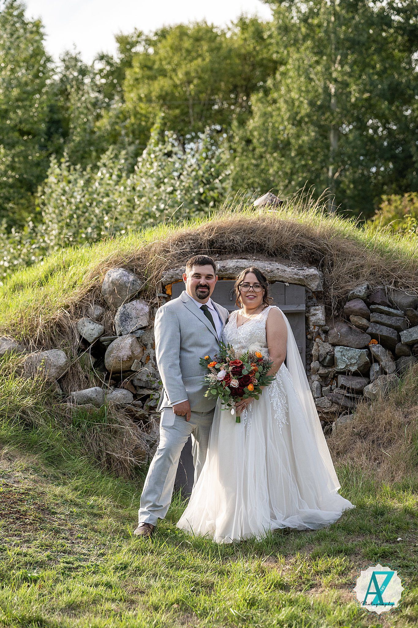 Newlyweds pose together near rustic stone wall in sunlit outdoor garden setting during wedding photoshoot.