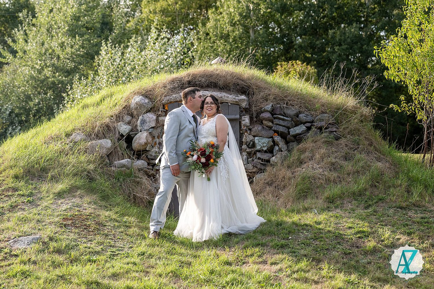 Wedding couple standing on a grassy hillside in formal attire with natural stone wall and greenery in background.