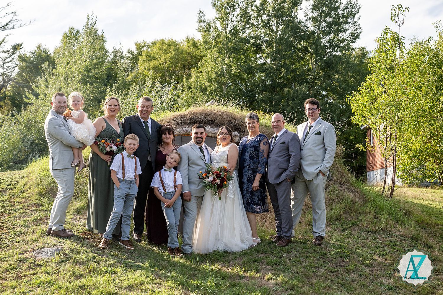 Wedding group photo with bridesmaids in white dresses and groomsmen in gray suits posed in a garden setting.