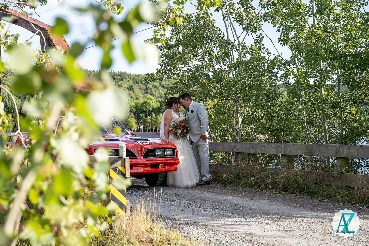 A couple shares an intimate moment near a red Mustang car on a tree-lined country path.