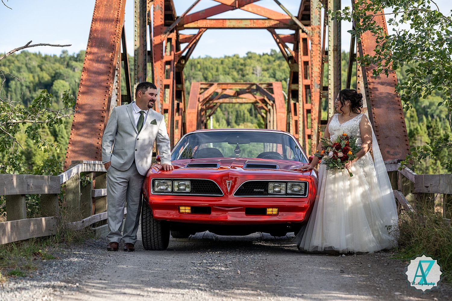 A classic red car parked under a wooden bridge creates a unique backdrop for a wedding photo session.