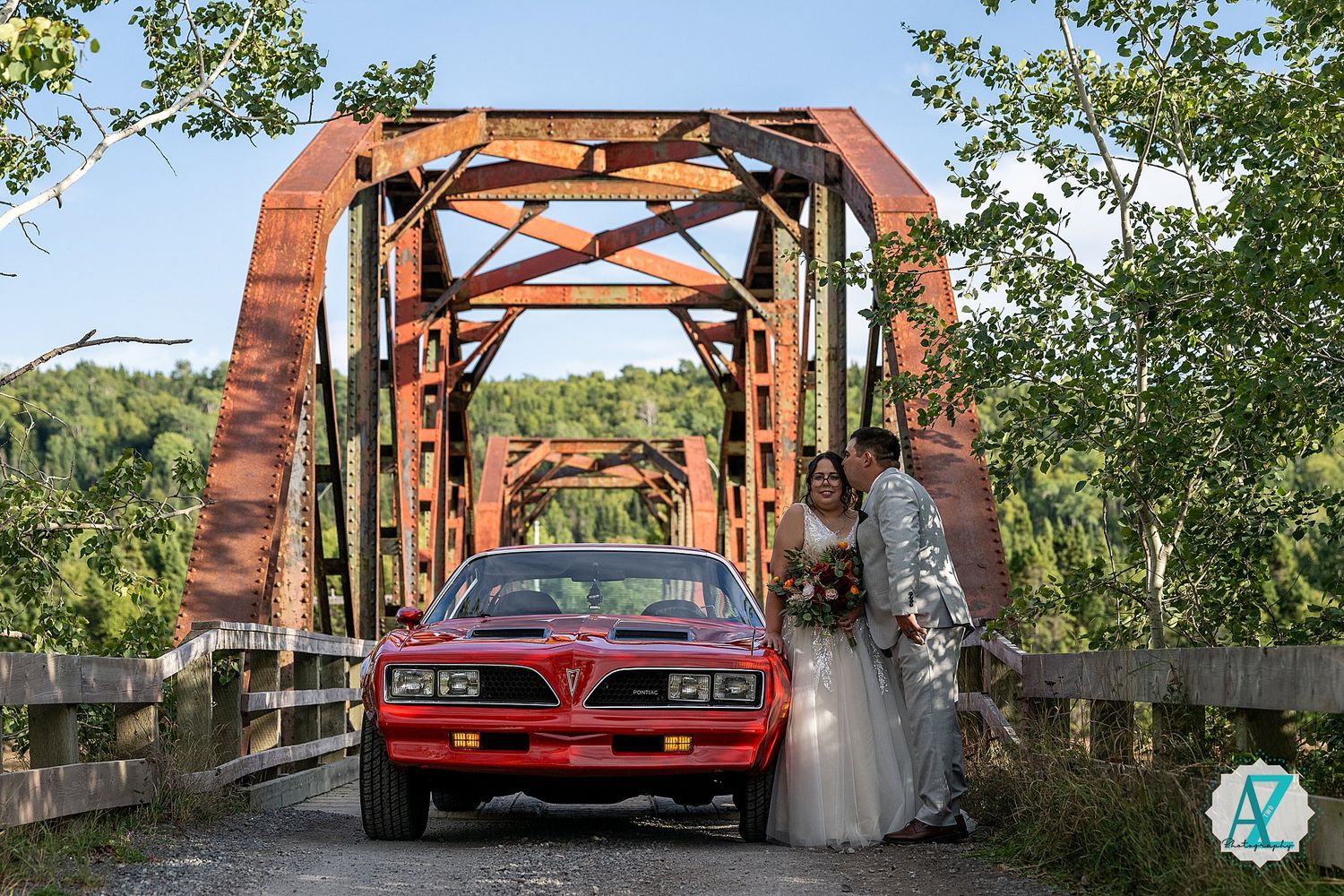 A red vintage GTO car photographed in front of a rustic wooden bridge with trees in the background.