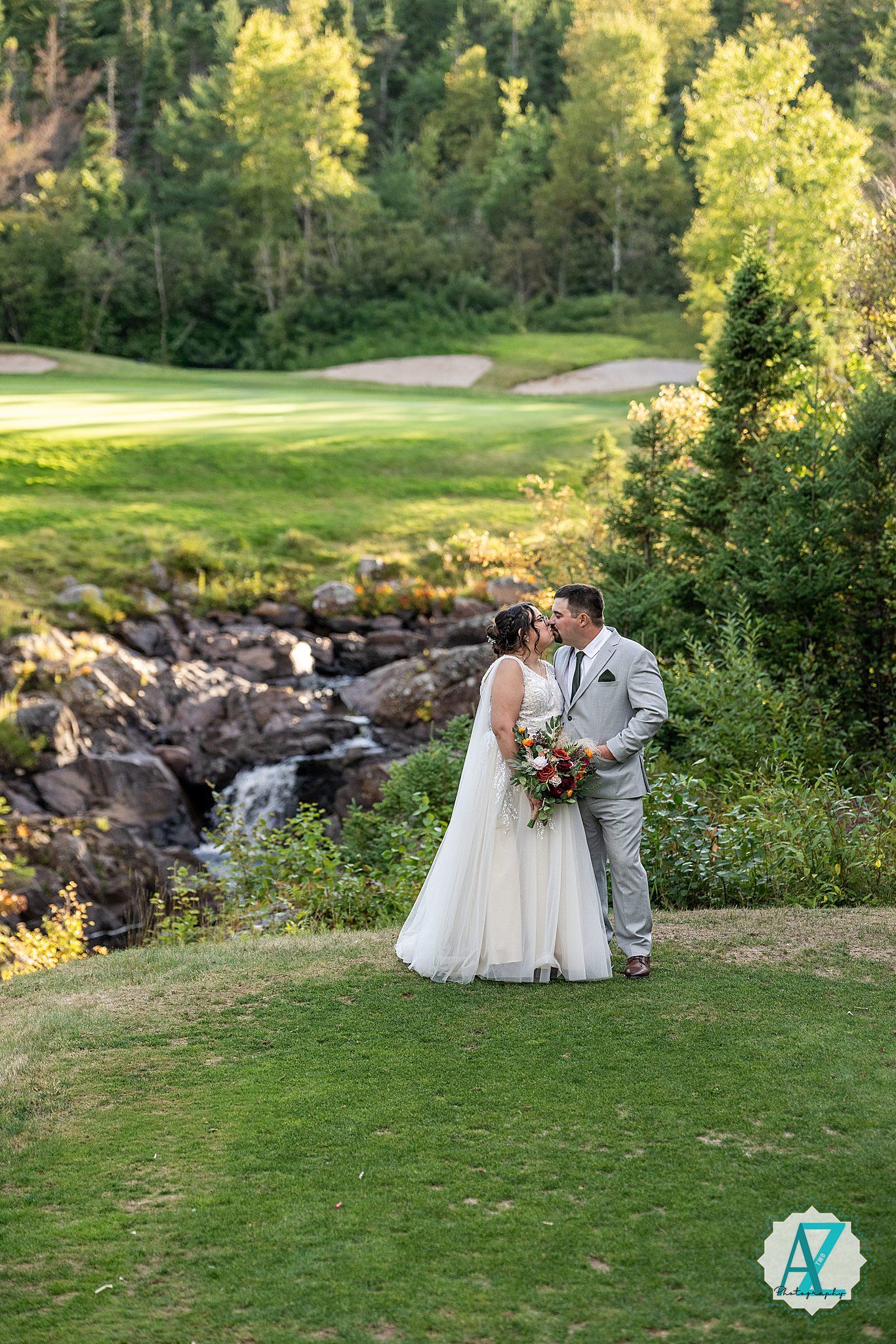 Wedding couple sitting on grassy hill with scenic waterfall and rocks in the background.