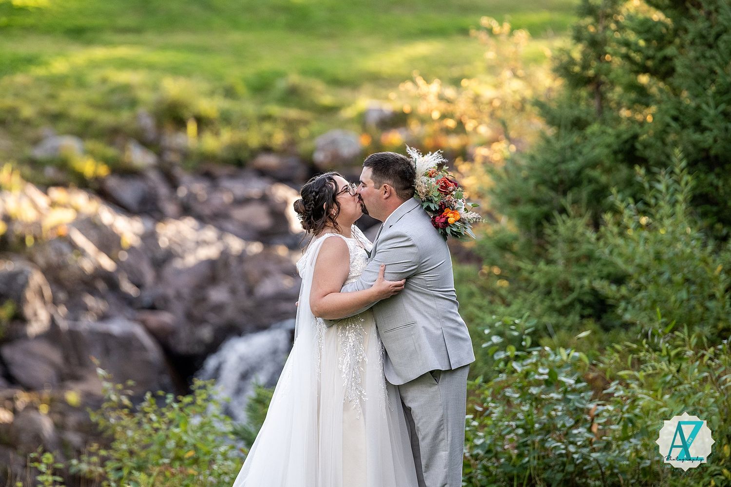 Two figures stand together in a romantic embrace among lush greenery and rocks on a sunny summer day.