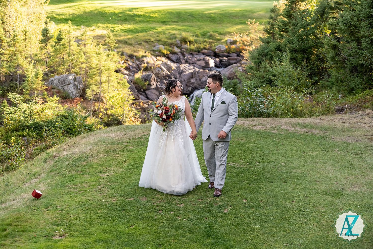 A couple poses together on a lush green hillside surrounded by scenic mountain views at an outdoor wedding celebration.
