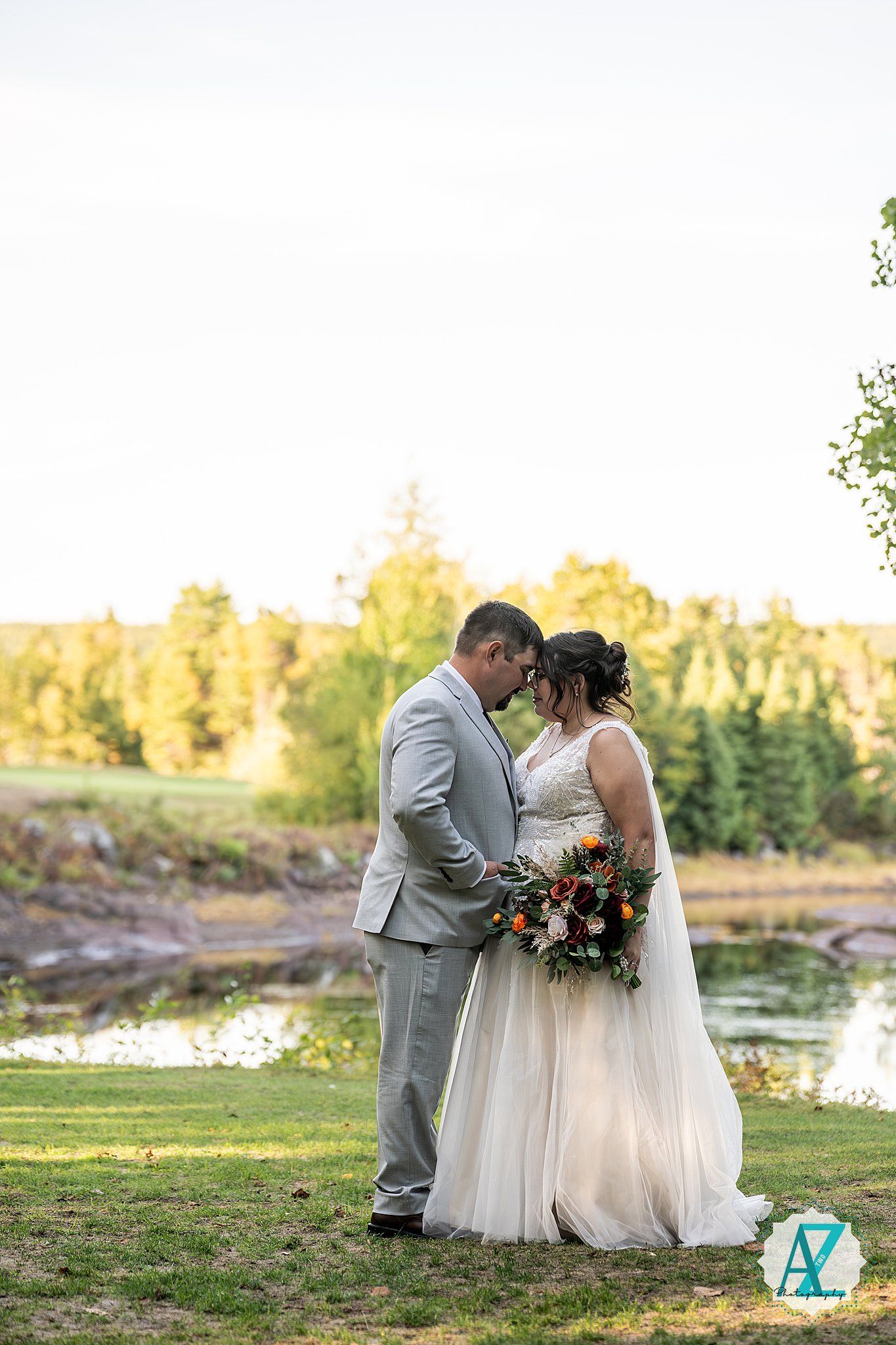 Sunset wedding portrait with couple sitting on grassy hill near waterfront with autumn colors.