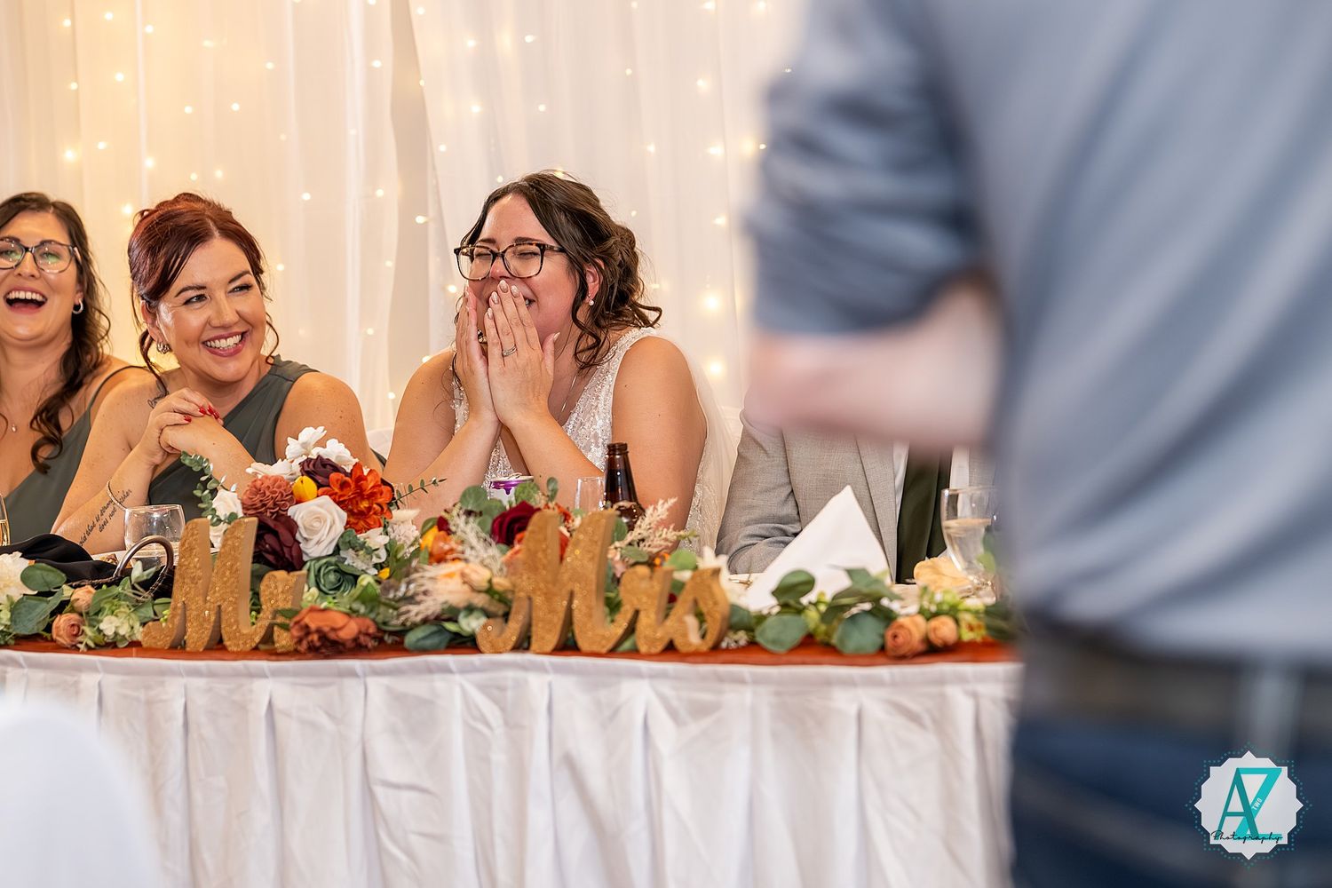 Wedding reception guests react with joy and laughter during speeches at a decorated banquet hall.