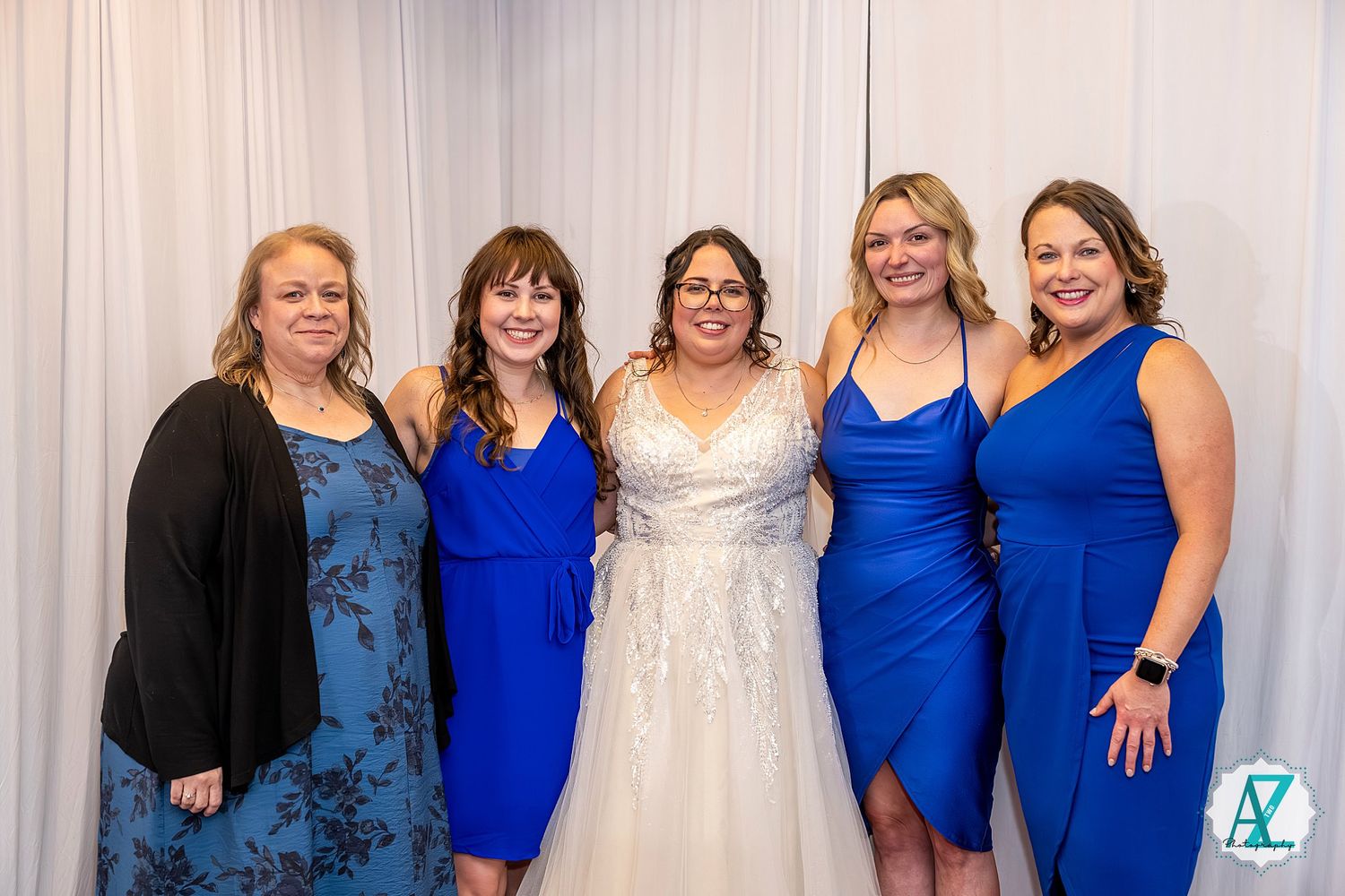 Wedding party members in coordinated royal blue dresses pose together against a white backdrop at formal event.