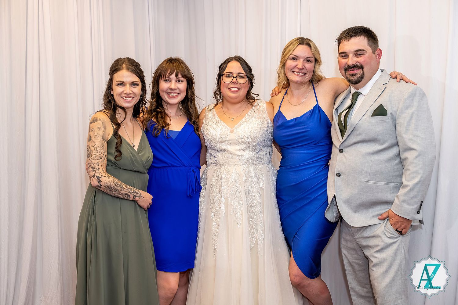 A group of people pose together at a wedding celebration with a white backdrop, featuring elegant blue and gray attire.