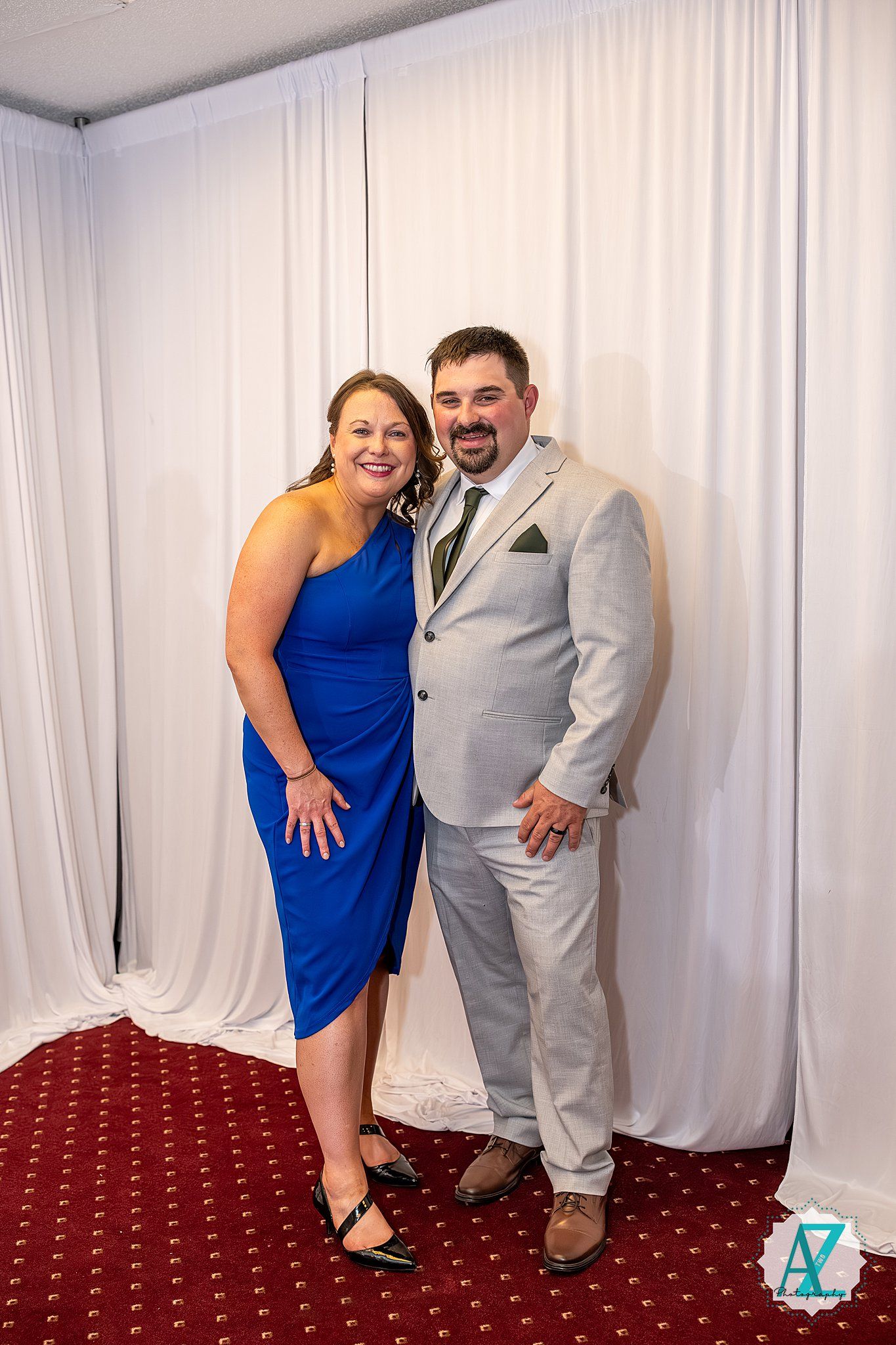 A couple in formal attire pose together against a white backdrop with a red carpeted floor.