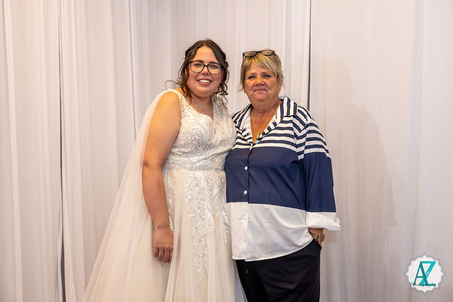 Two people pose together at a formal event with white backdrop, one wearing a white dress and one in blue striped top.