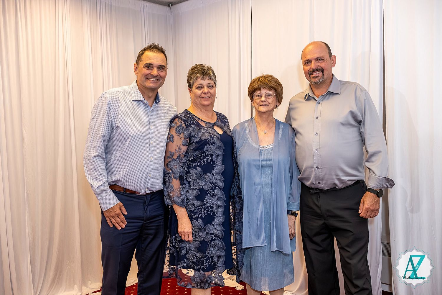 Group of four adults in formal and casual attire posing together against a white curtain backdrop at an event.