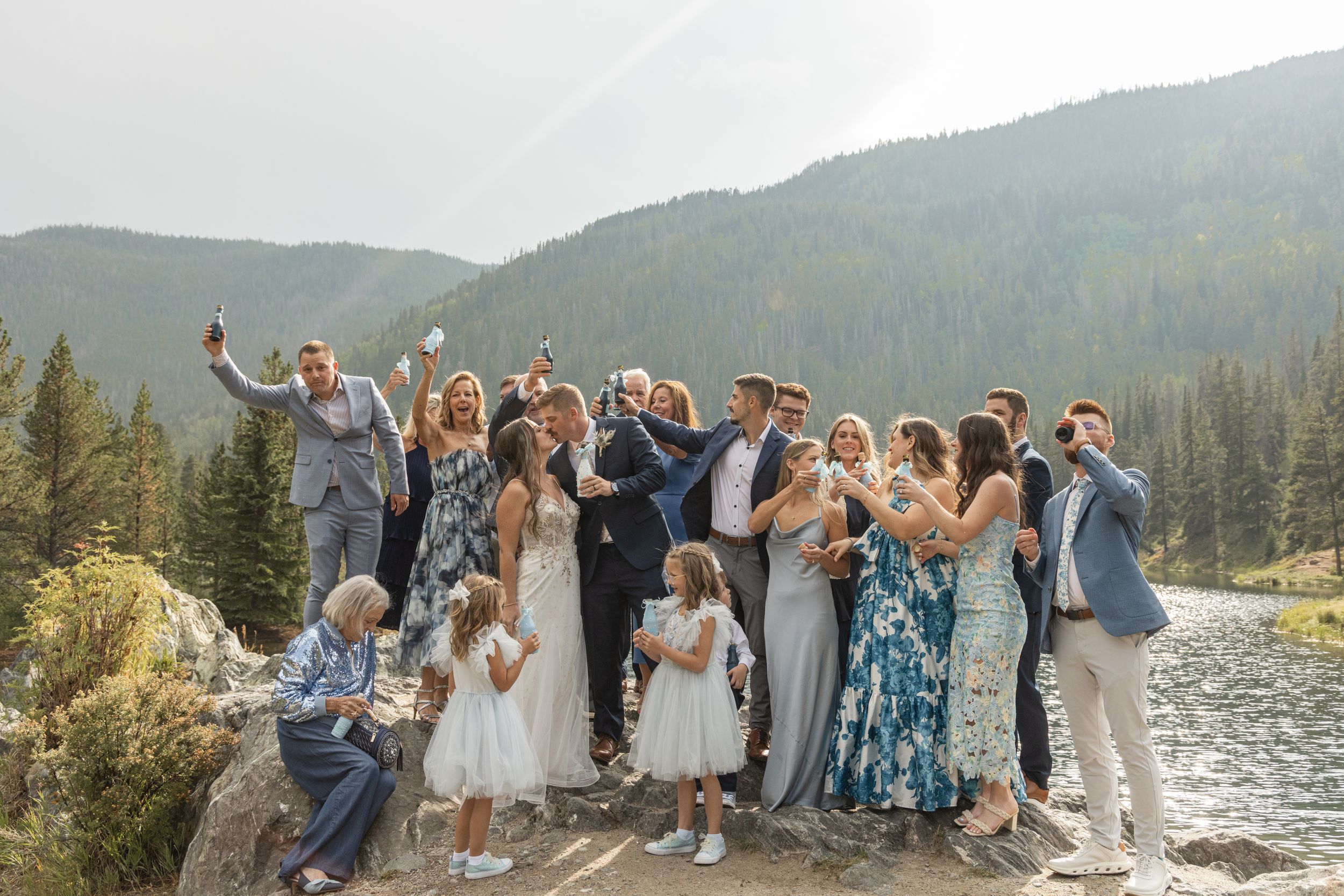 Wedding party gathers on a rocky beach with mountain views and pine trees creating a scenic backdrop in nature.