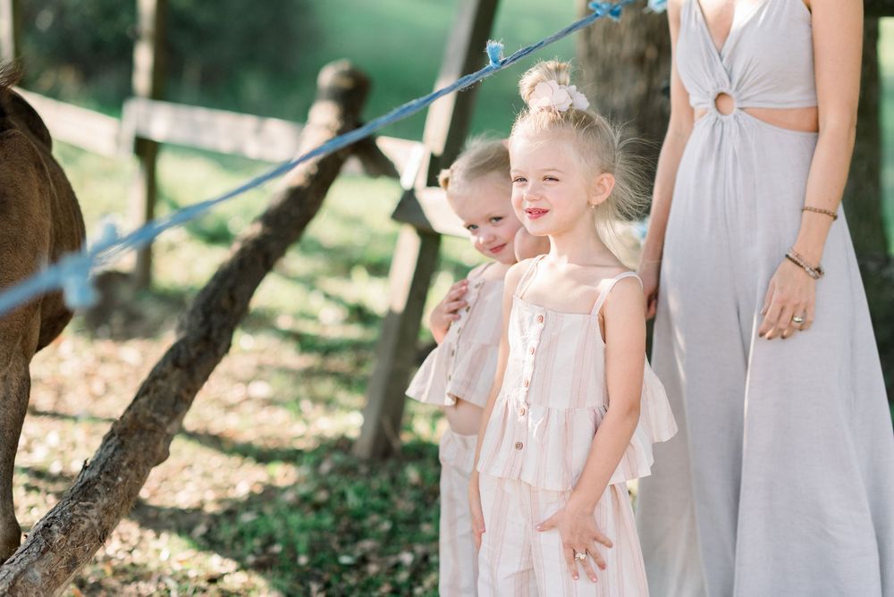 Group wearing light colored dresses stands together under trees in a natural setting.