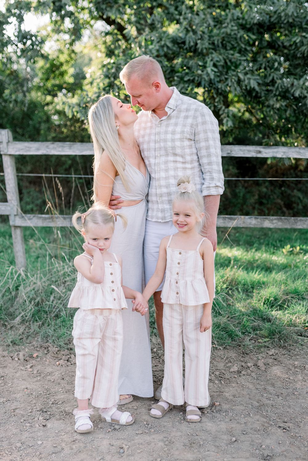 Family in coordinating neutral outfits poses together near a wooden fence in a rural setting.