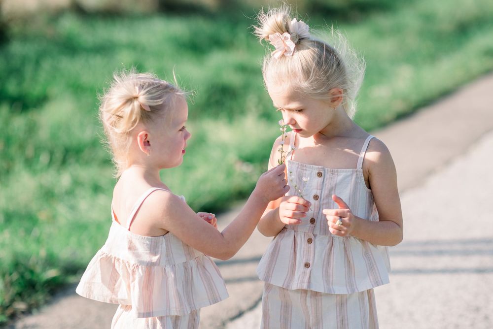 Two children in matching light-colored dresses interact playfully outdoors.