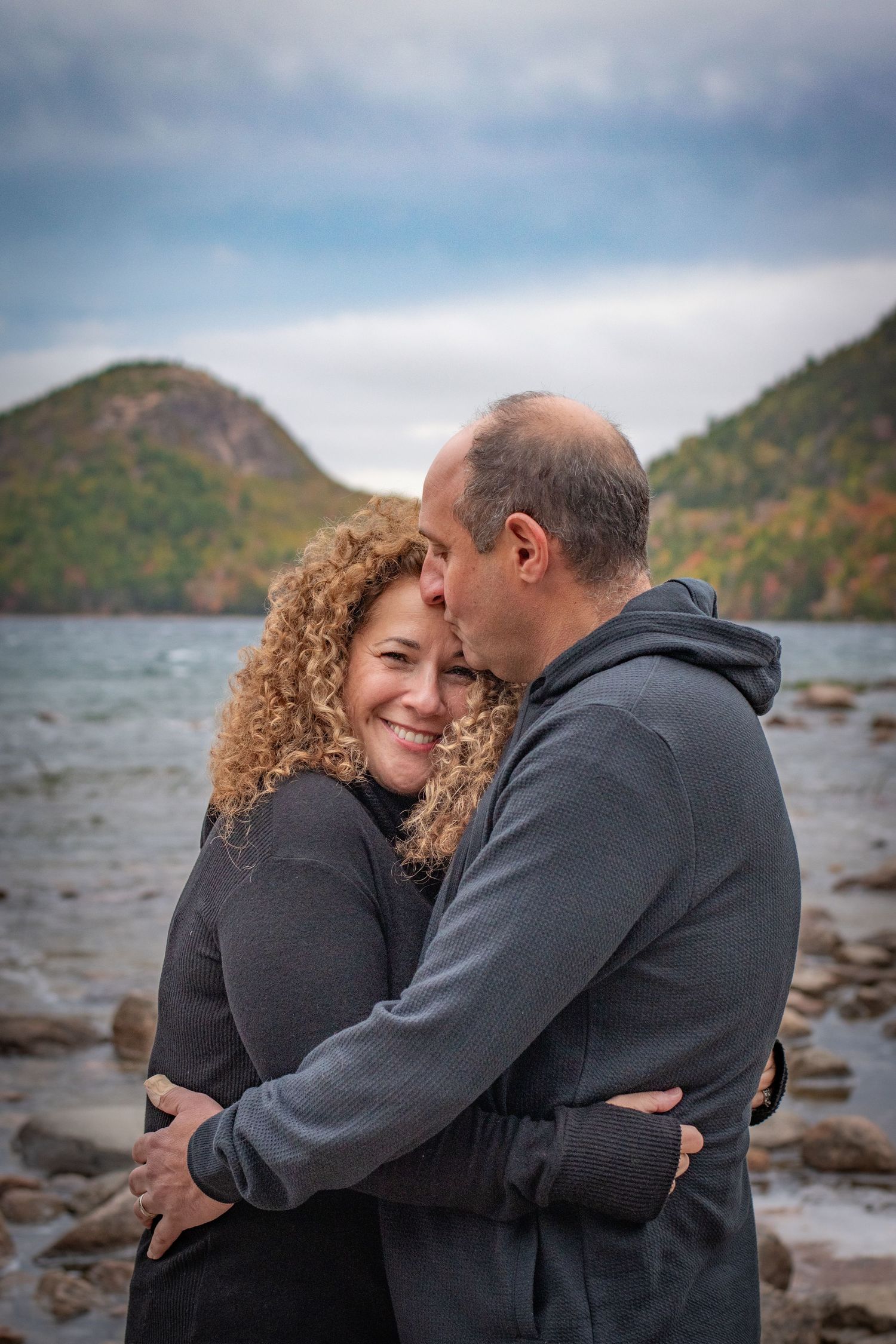 Romantic couple embracing and kissing by rocky lakeside with mountain backdrop on cloudy day. Photo by Xenophon Photography in Maine.