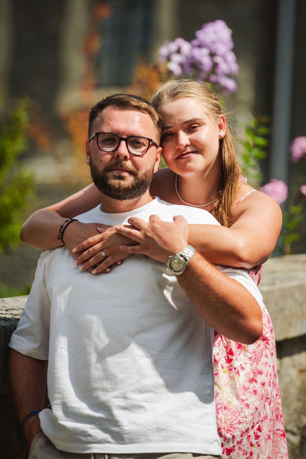 A couple embraces outdoors with pink flowers in the background during a romantic photoshoot.
