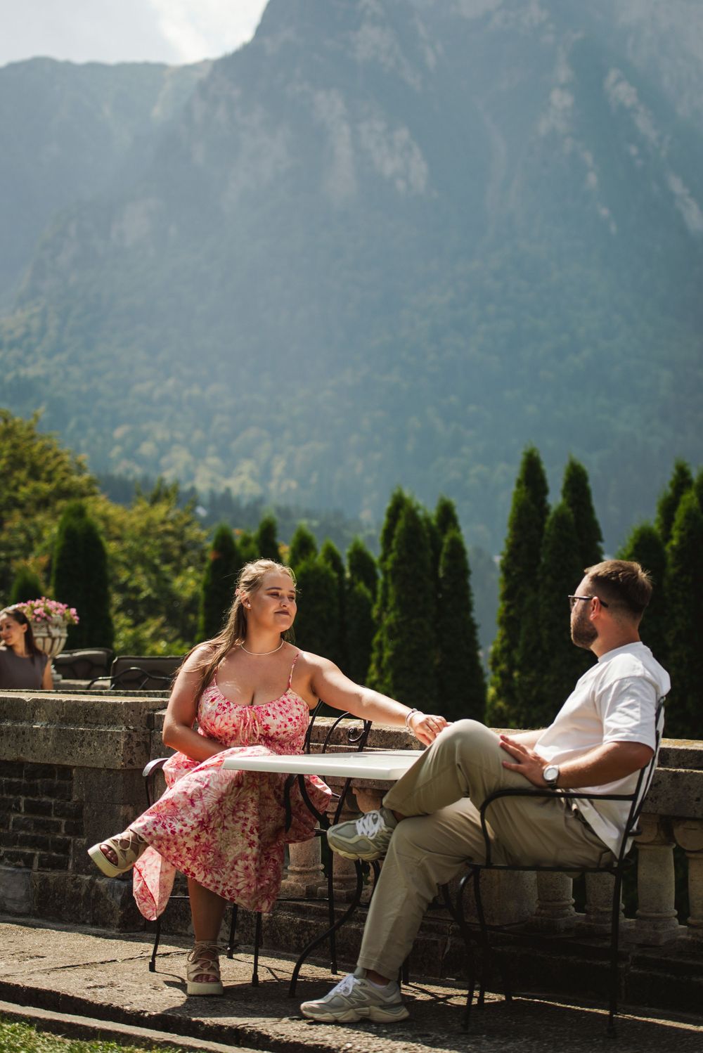 A couple relaxes on benches overlooking a scenic mountain landscape on a sunny day.
