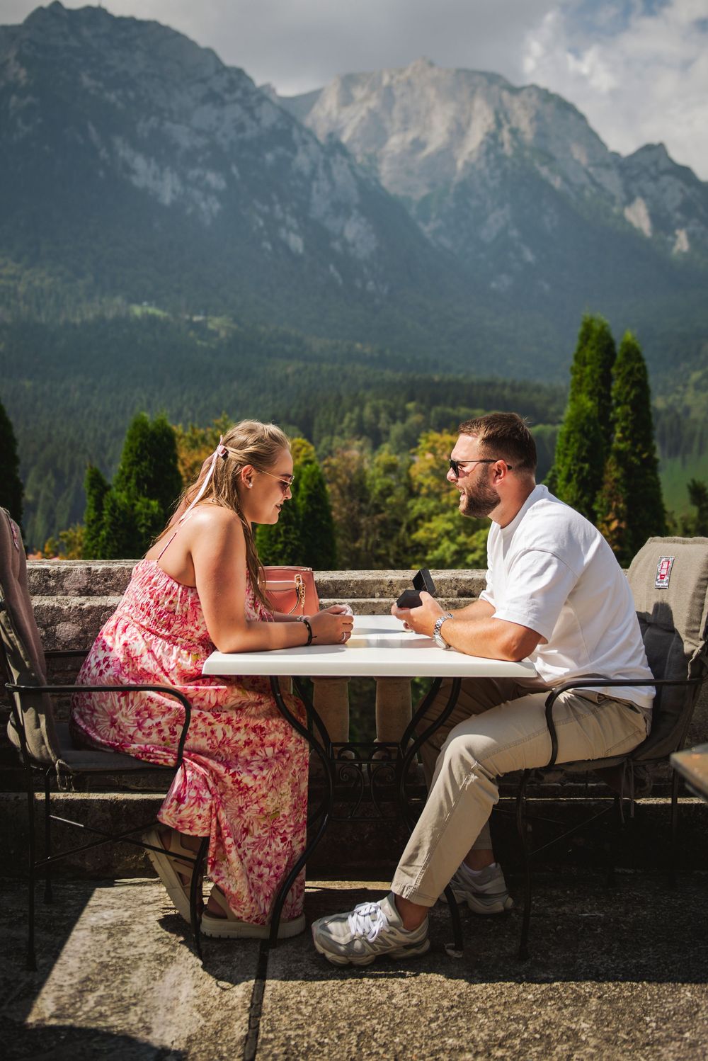 A couple sits at an outdoor table enjoying drinks with dramatic mountain peaks rising in the background.