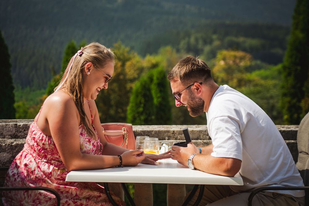 A couple sits together at an outdoor table in a mountain setting, looking down at papers or books on a sunny day.