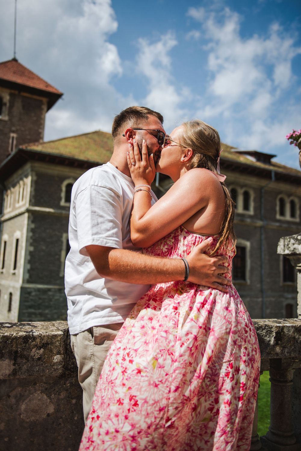 A couple shares a romantic kiss against a stone wall backdrop with a historic building in the background.