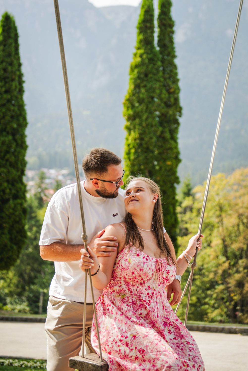 A couple shares a romantic moment on a swing with scenic mountain views and tall cypress trees in the background.