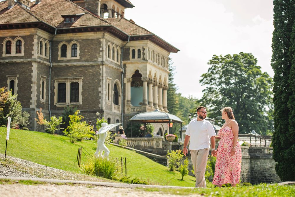 A couple walks together on the grounds of a historic stone mansion on a sunny summer day.